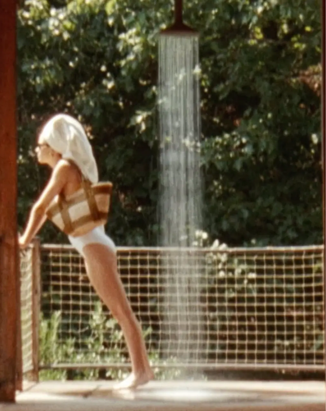 Woman in white carrying a woven tan bag beside an outdoor shower